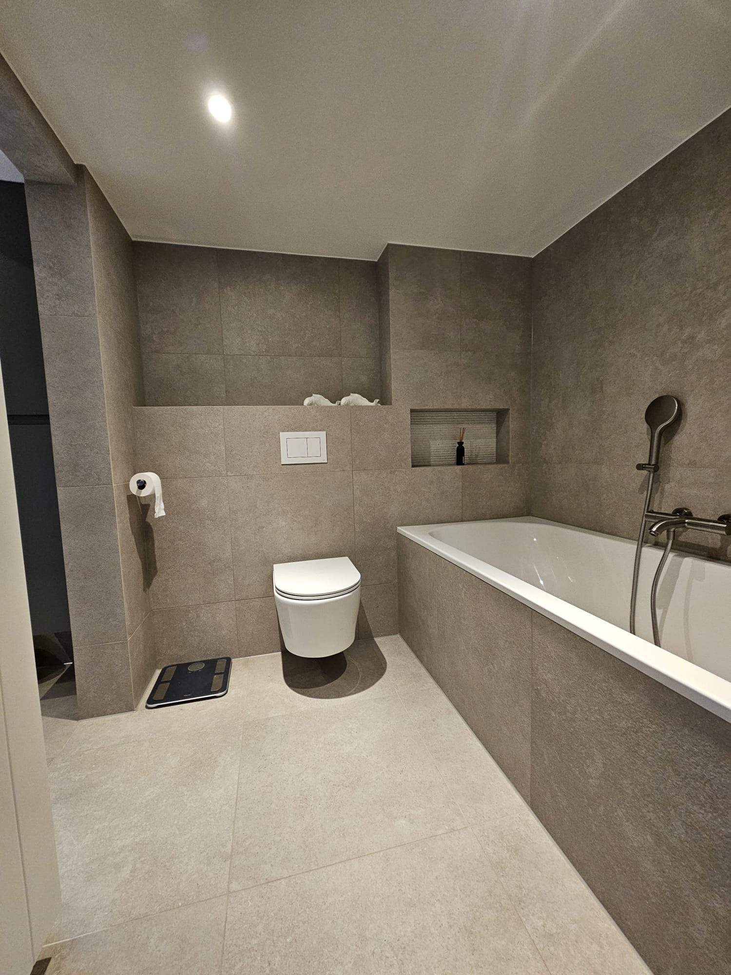 Afgewerkte badkamer in warme betonlook met betegelde badomkasting, nis en zwevend toilet